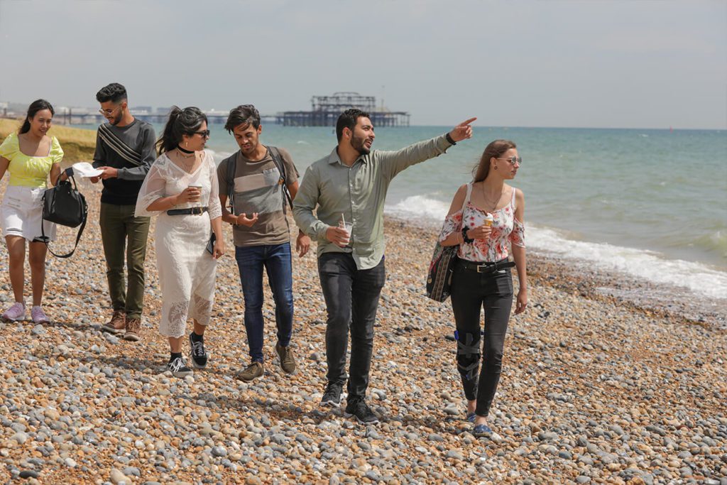 A group of students walk along Brighton beach, pointing out to sea