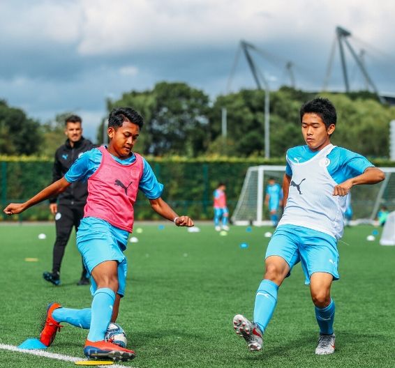 Two teenage boys playing football as Manchester City Academy coach looks on in background
