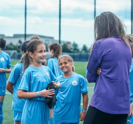 Girls in full Manchester City kit talking with female coaching staff