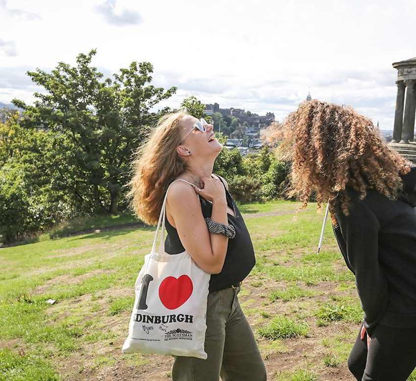 Students laughing in a park, holding an Edinburgh bag.