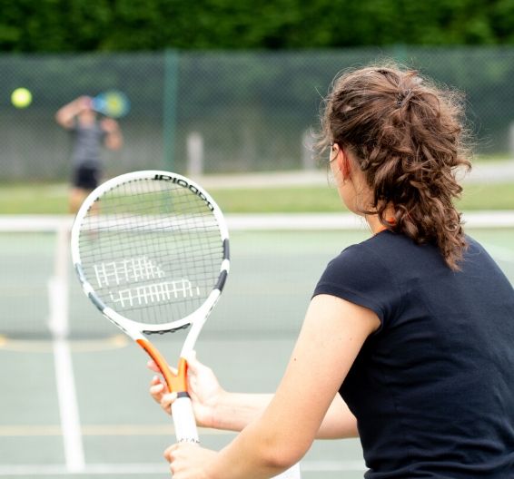 A girl holds a tennis racquet up, preparing to volley the ball served by her opponent
